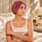 A woman wearing a dusty rose turban-style hair cap sits on a bed with shiny satin bedding in a warm, elegant bedroom setting.