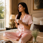 Woman in pink pajamas sitting on a bed holding a cup, with a tray of food and a magazine beside her.